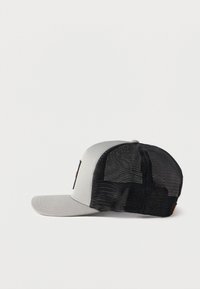 Casquette trucker grise à l'avant avec un filet noir à l'arrière et une visière plate, vue de profil sur un fond blanc.
