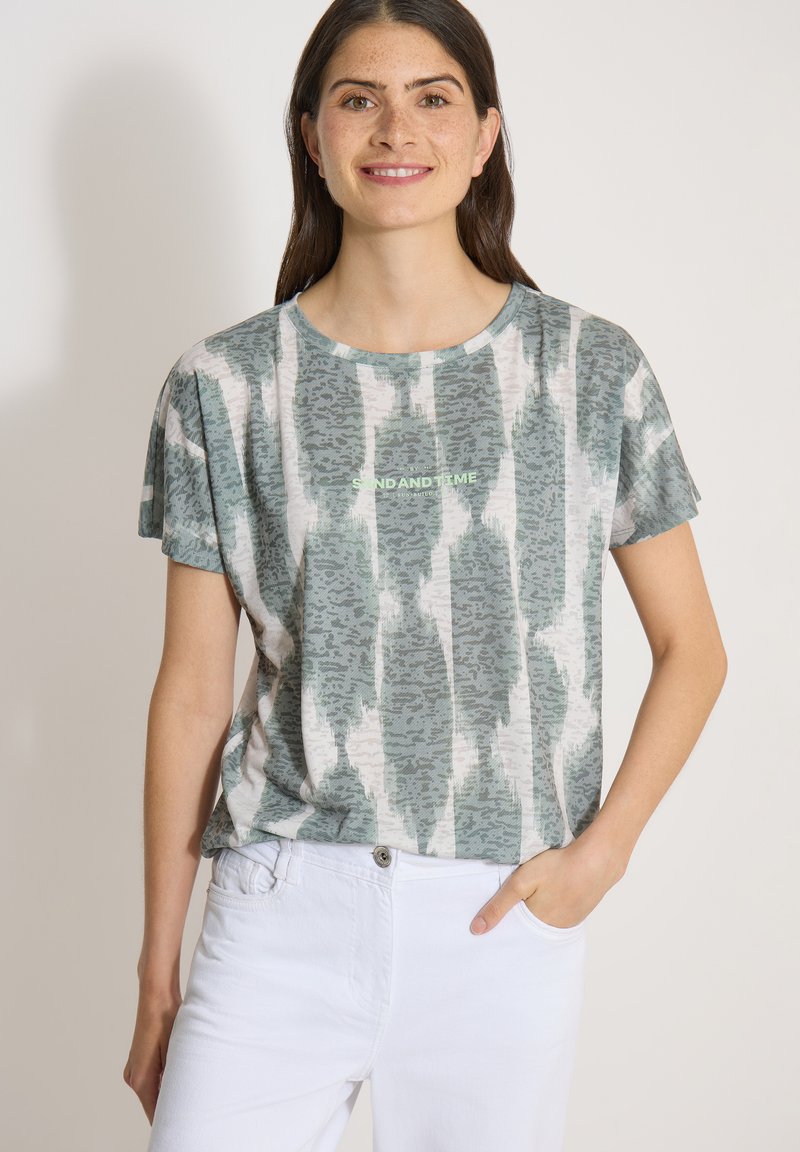 Femme aux longs cheveux bruns portant un t-shirt vert et blanc à motifs et un pantalon blanc, souriant avec une main dans la poche.