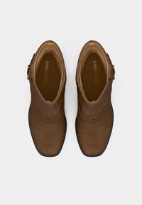 Brūni zamšādas potīšu zābaki ar noapaļotu purngalu, rotāti ar apkārtējiem elementiem un dekoratīvu zelta toņa sprādzi ārējā pusē.