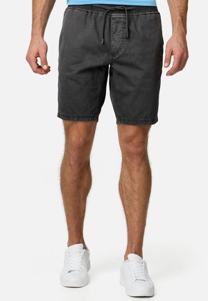 Homme portant un short noir avec cordon, des baskets blanches et une chemise bleu clair, debout devant un fond uni.