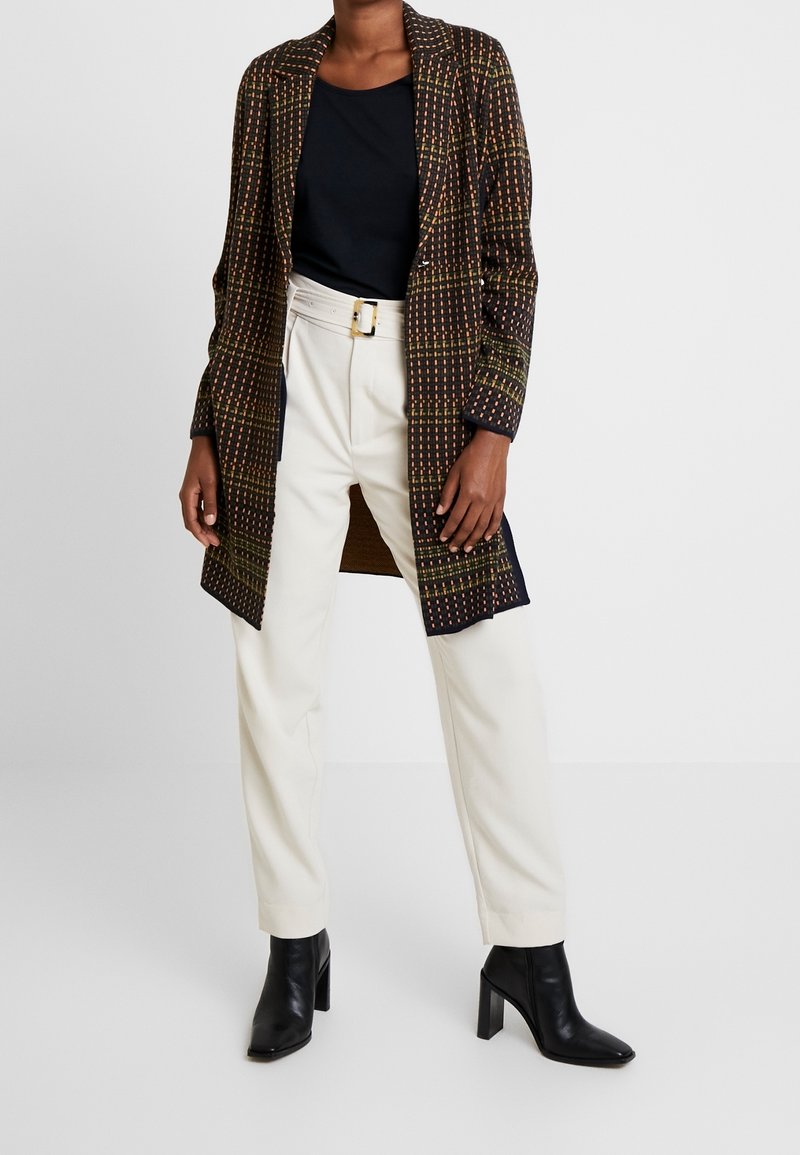 Blazer lungo in tessuto scuro con un design a motivi che presenta accenti arancioni e verdi. Abbinato a pantaloni bianchi e stivaletti neri con tacco.