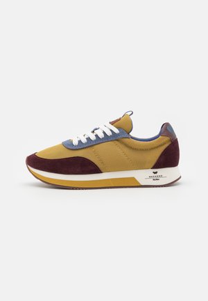 Niski sneaker z żółtą siateczką, bordowymi zamszowymi akcentami, niebieskim panelem z dziurkami na sznurowadła, białymi sznurowadłami oraz białą i żółtą podeszwą na białym tle.