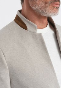 Szary, strukturalny żakiet z kołnierzem stojącym i brązowymi, zamszowymi detalami. Noszony na białej koszuli, prezentujący elegancki, nowoczesny design.