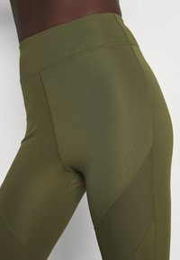Olijfgroene leggings van rekbaar materiaal met een gladde textuur. Voorzien van contrasterende meshpanelen en platte naden voor comfort.