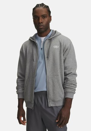 Man met gevlochten haar die een lichtgrijze zip-up hoodie draagt over een blauw shirt met een kwart-rits en donkergrijze broek tegen een witte achtergrond.