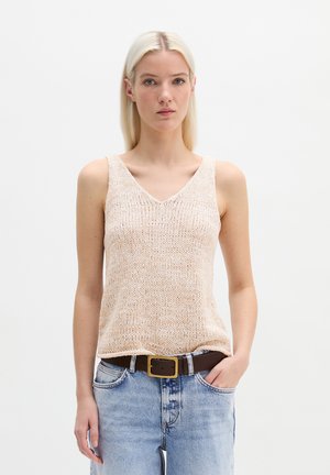 Blonde Frau trägt ein ärmelloses, beiges Stricktop, das in eine hellblaue Jeans mit einem dunkelbraunen Gürtel gesteckt ist, und steht vor einem einfarbigen weißen Hintergrund.