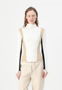 Garu piedurkņu zip-up tops baltā, bēšā un melnā krāsā. Tam ir piegulīga forma, augsts apkaklis un pirkstu caurumi pie piedurknēm. Gluda, elastīga auduma.