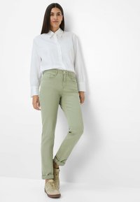 Hellgrüne Baumwollhose im Slim-Fit-Schnitt, kombiniert mit einem weißen Hemd mit Kragen. Sie verfügt über hochgerollte Säume und hellbraune Sneakers.