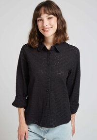 Chemise noire à manches longues avec un design texturé en dentelle avec des motifs floraux. Manches longues avec poignets roulés. Coupe décontractée.