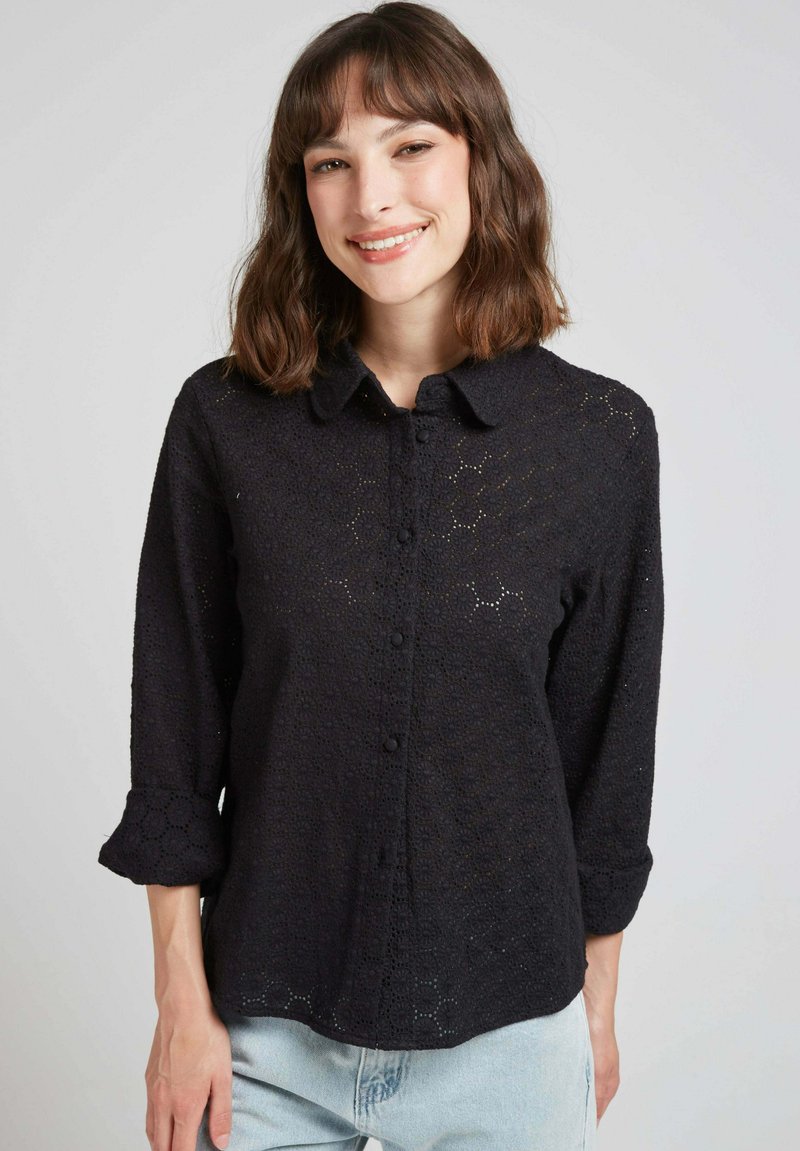 Chemise noire à manches longues avec un design texturé en dentelle avec des motifs floraux. Manches longues avec poignets roulés. Coupe décontractée.