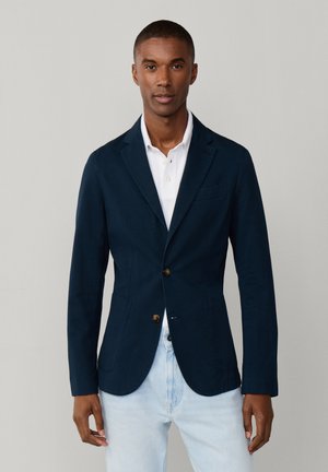 Hombre vestido con un blazer azul marino sobre una camisa blanca y vaqueros azul claro, posando contra un fondo liso de color gris claro.