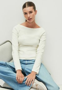 Biele priliehavé tričko s dlhým rukávom a širokým výstrihom, skombinované s svetlomodrými džínami. Modelka sedí uvoľnene na šedej stoličke.