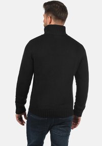 Schwarzer gestrickter Pullover mit hohem Kragen, gerippten Bündchen und Saum, mit strukturierter Oberfläche. Getragen zu blauen Jeans.