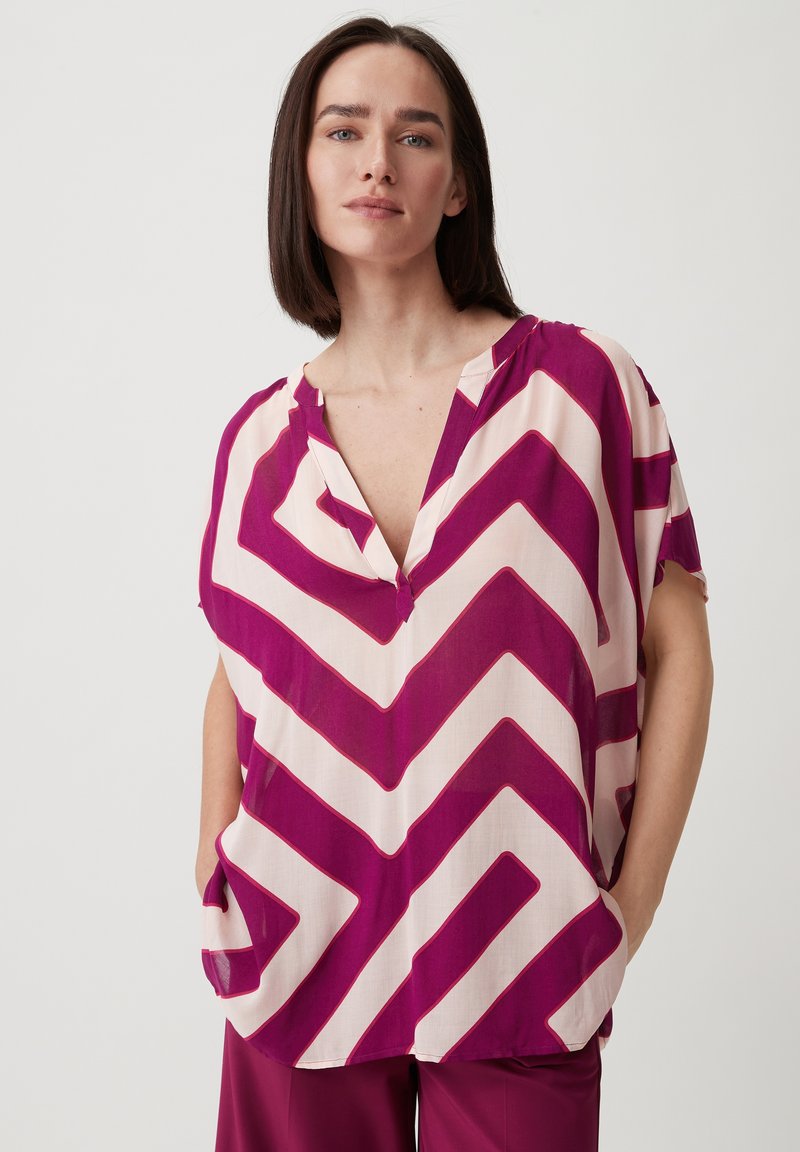 Bluzka typu v-neck z lekkiego materiału w zygzakowaty wzór w fuksjowym i kremowym kolorze. Krótkie rękawy i boczne kieszenie zwiększają funkcjonalność.