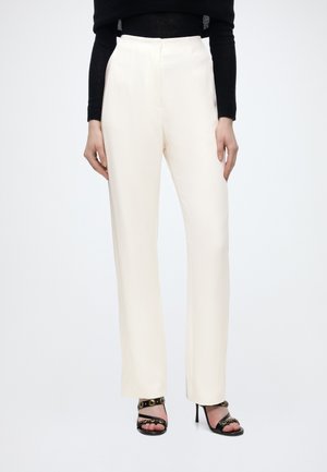Vrouw draagt witte, recht gesneden broek met hoge taille, gecombineerd met zwarte hakken en een zwarte top met lange mouwen, staand tegen een witte achtergrond.