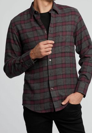 Mężczyzna ubrany w szarą i bordową koszulę w kratę na guziki, noszoną na czarnej koszulce, jedną ręką poprawia koszulę, a drugą ma w kieszeni.