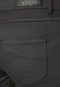 Närbild av svart denimtyg, med en bakficka som har distinkta sömmar och en liten läderetikett med texten 'SISLEY' vid midjan.