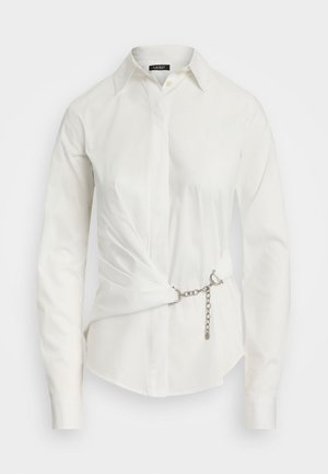 Chemise blanche à manches longues et col, avec un détail de devant torsadé maintenu par une ceinture en chaîne argentée.