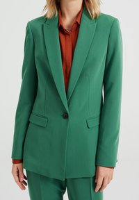 Zielona marynarka o dopasowanym kroju, z pojedynczym czarnym guzikiem i dwoma przednimi kieszeniami. Noszona na rdzawym kolorze koszuli, tworząc kontrastujący wygląd.