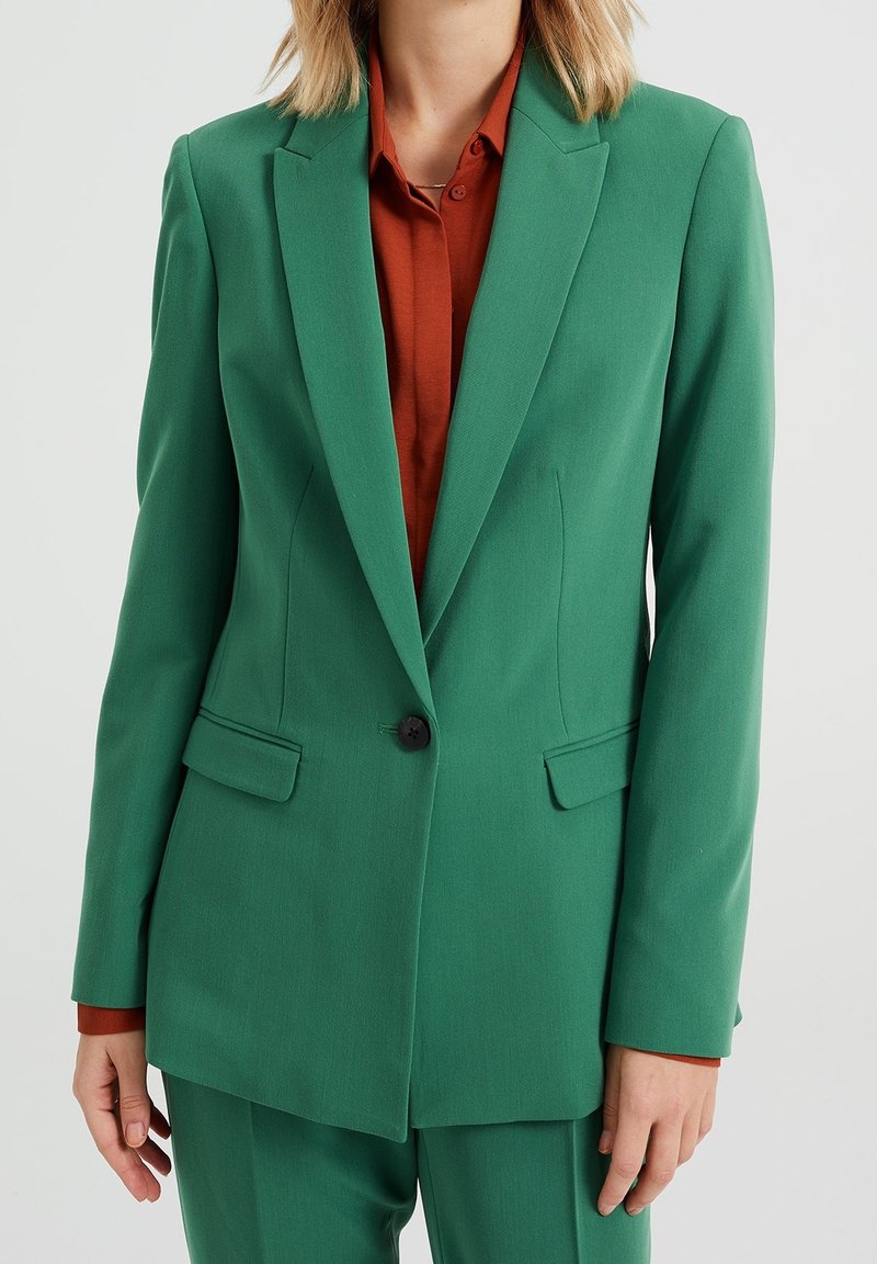 Zielona marynarka o dopasowanym kroju, z pojedynczym czarnym guzikiem i dwoma przednimi kieszeniami. Noszona na rdzawym kolorze koszuli, tworząc kontrastujący wygląd.