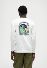 Langärmliges weißes Hemd mit buntem Grafikdesign eines Skeletts, das auf einer Welle surft, auf dem Rücken, mit fettgedrucktem Text "RIP CURL" darüber.