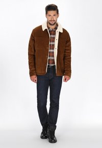 Braune Cord-Jacke mit einem weißen, mit Fell gefütterten Kragen, ausgestattet mit zwei Brusttaschen. Kombiniert mit einem karierten Hemd, dunklen Jeans und schwarzen Stiefeln.