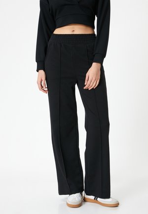 Zwarte wijdvallende sweatpants met een elastische tailleband, met een gladde textuur en gestructureerde naden, gedragen met witte sneakers.