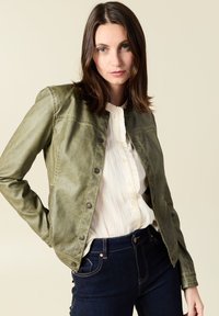 Giacca in pelle verde oliva con chiusura a bottoni, caratterizzata da un collo classico e una texture liscia, indossata su una blusa bianca con volant e jeans scuri.