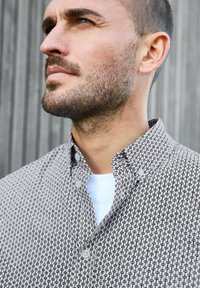 Camisa de botones con un patrón geométrico en blanco y negro, que cuenta con un cuello puntiagudo y botones en contraste. Camiseta blanca visible por debajo.