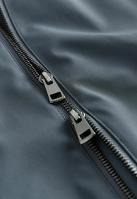 Tumši zils audums ar rāvējslēdzēju, gluda tekstūra, ar diviem sudraba un melniem rāvējslēdzējiem, redzamas detaļas šuvēs.