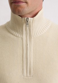 Man die een roomkleurige geribbelde gebreide trui draagt met een zilveren ritssluiting, deels dichtgeritst tot aan de nek, waarbij kin en onderkant van het gezicht zichtbaar zijn.