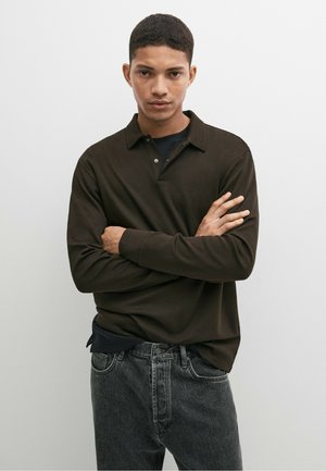 Junger Mann mit kurzen Haaren, der ein dunkelbraunes langärmeliges Poloshirt und graue Jeans trägt und mit verschränkten Armen vor einem schlichten Hintergrund steht.