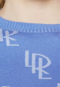 Maglione blu in maglia con scollo a costine e un motivo di lettere bianche distribuite nel tessuto, creando contrasto e texture.