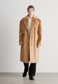 Garšs, pogām aizdara mētelis viegli brūnā mākslīgā vilnā, ar plašu apkakli, teksturētu apdari un sānu kabatām, valkāts virs ribotā krekla.