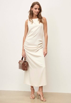 Femme portant une robe midi crème sans manches et des talons dorés à fines lanières, tenant un sac à main en cuir marron, debout contre un mur uni.