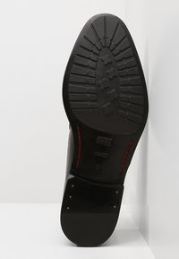 Semelle de chaussure en cuir noir avec un motif texturé, présentant une couture rouge contrastante et de petites clous en métal près de la zone du talon.