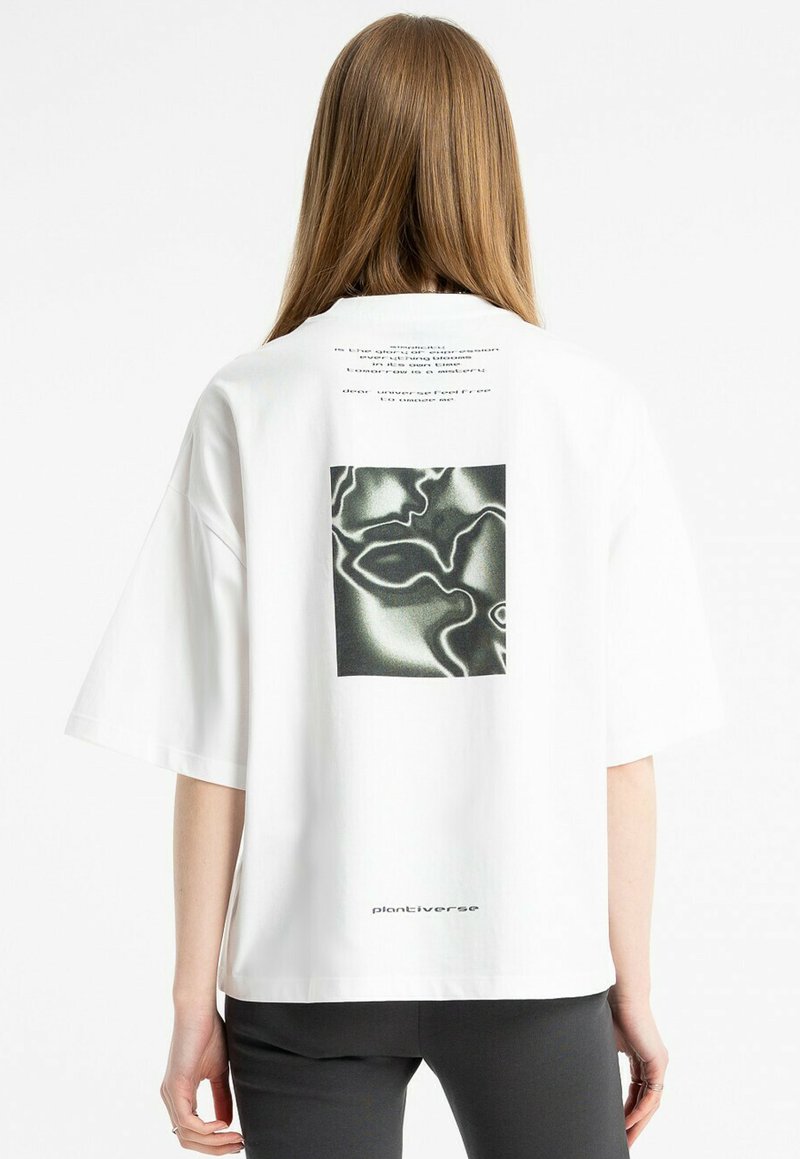 Osoba z długimi włosami nosi w oversize'owej, białej koszulce z abstrakcyjną czarno-białą grafiką i tekstem na plecach, do tego czarne spodnie.