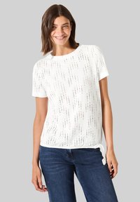 T-shirt bianco a maniche corte con motivo perforato, scollatura rotonda e dettaglio con fiocco laterale. Abbinato a jeans blu. Texture liscia.