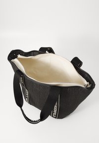 Black woven tote bag with a zip closure and cream interior. Features black straps with white text. Structured shape with a textured surface.