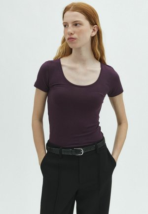 Jeune femme aux longs cheveux roux portant un t-shirt moulant violet foncé à col rond et un pantalon noir avec une ceinture noire, mains dans les poches, regardant vers la gauche.