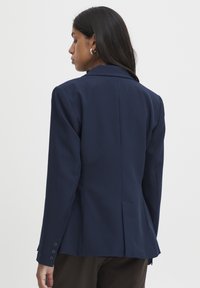 Blazer bleu marine ajusté, avec des revers à cran et des manches longues. Présente une fente unique à l'arrière et des poignets boutonnés. Texture de tissu lisse.