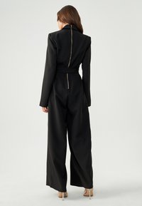 Zwarte jumpsuit met een aansluitende bodice en wijde pijpen. Beschikt over een hoge kraag en een gouden rits aan de achterkant. Glad materiaal met een ceintuur in de taille.