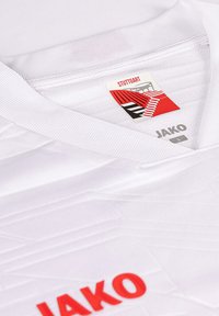 Maglia sportiva bianca con design a scollo a V, caratterizzata da strisce testurizzate e un logo rosso "Jako", con un'etichetta che mostra "Stuttgart" sotto.