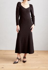 Svart texturerad långärmad klänning med fyrkantig halsringning, kompletterad med svarta spetsiga klackar med guld accenter.