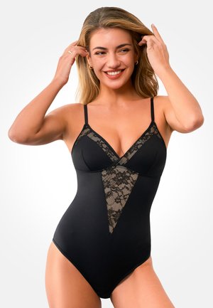 Uśmiechnięta kobieta poprawiająca włosy, ubrana w czarny body z koronkowymi zdobieniami na piersiach i przodzie, na białym tle.