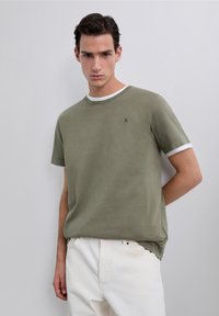 Jonge man die gelaagde olijfgroene en witte korte mouw shirts draagt met witte broek, staand tegen een effen lichtgrijze achtergrond.