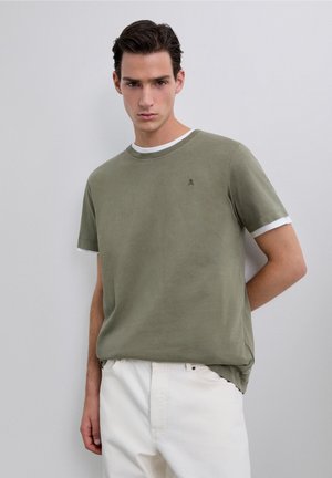 Jonge man die gelaagde olijfgroene en witte korte mouw shirts draagt met witte broek, staand tegen een effen lichtgrijze achtergrond.