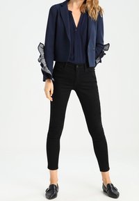 Kvinna i en stilren marinblå kavaj med volangärmar, matchad med en mörk topp och svarta jeans, poserar självsäkert i en neutral miljö.