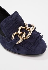 Mocassin en suède bleu marine avec détail à franges et grande chaîne dorée en embellissement sur le dessus, forme arrondie à l'avant.