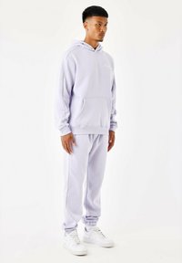 Lichtpaarse hoodie en bijpassende joggingsbroek van zacht materiaal, met een voorzak, ribgebreide manchetten en details bij de enkels. Witte sneakers.